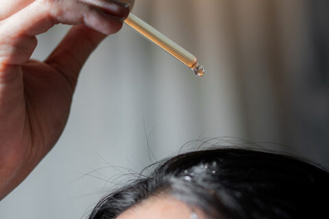 A hand holds a glass dropper dispensing liquid onto the scalp